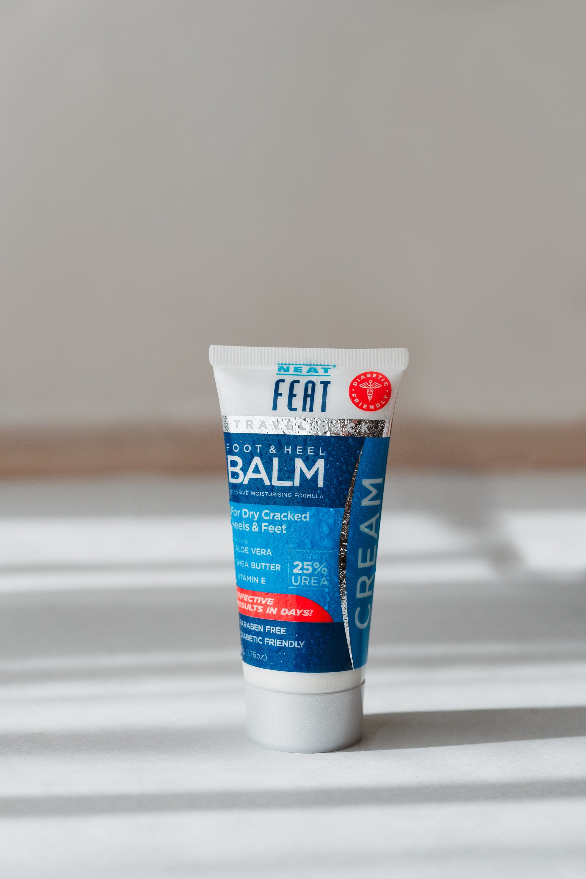 Front view of Neat Feat Foot & Heel Balm Travel Pack 50g tube, displayed on a grey surface with shadows and beads of water on the tube, featuring a grey cap and a blue label.
