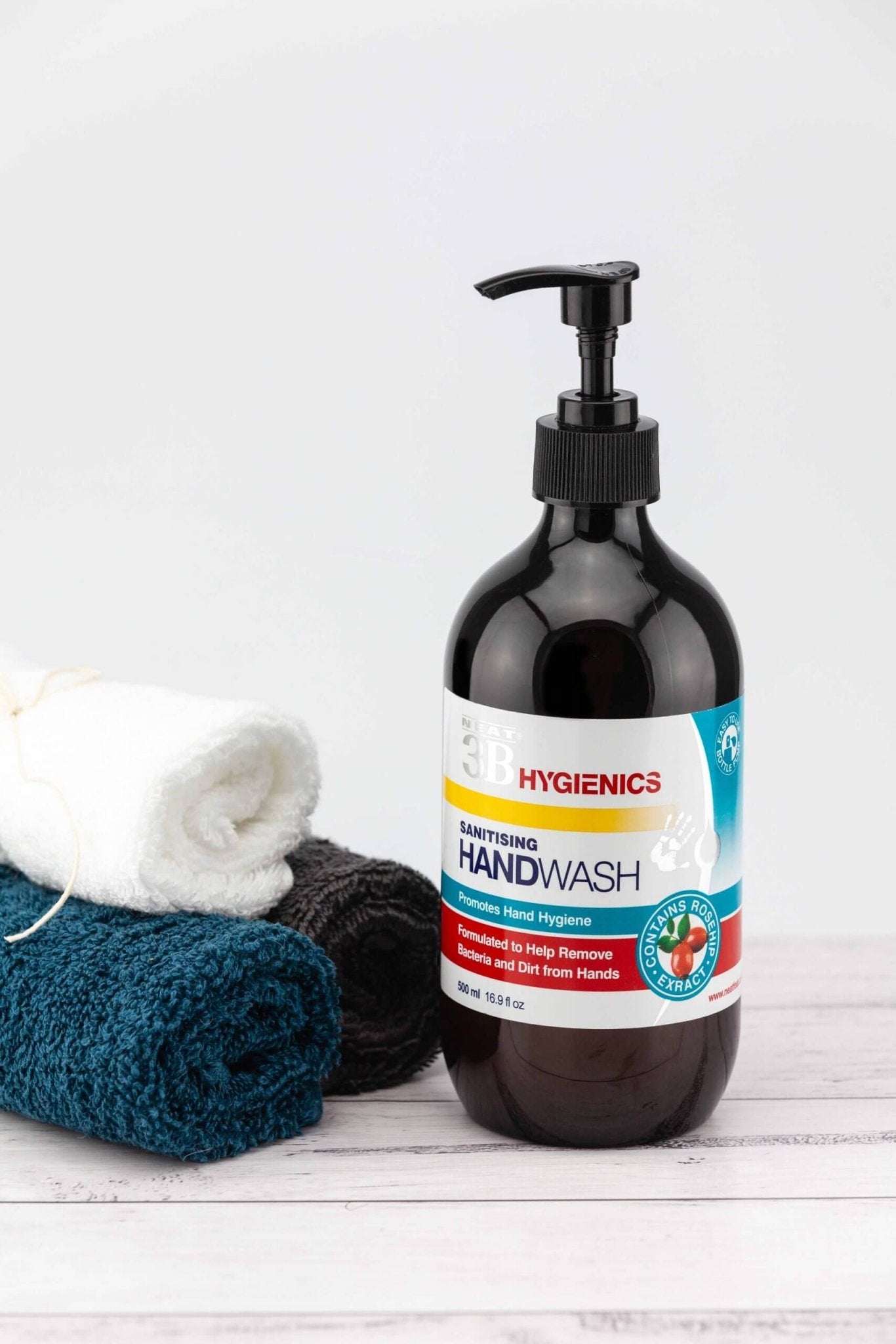 Neat Feat Hygienics Sanitising Handwash, front-on in a dark brown bottle with a black cap, featuring a white label with blue and red accents, displayed on a natural white table next to three small towels.