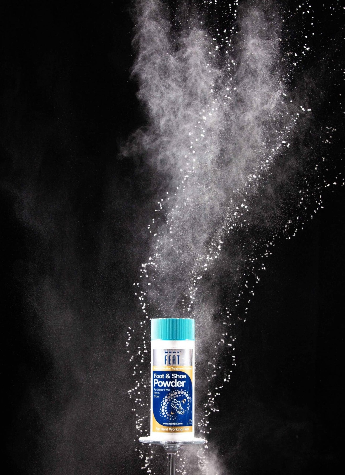 Neat Feat Foot & Shoe Powder displayed on a small table with a black background, featuring an explosion of powder behind the product.